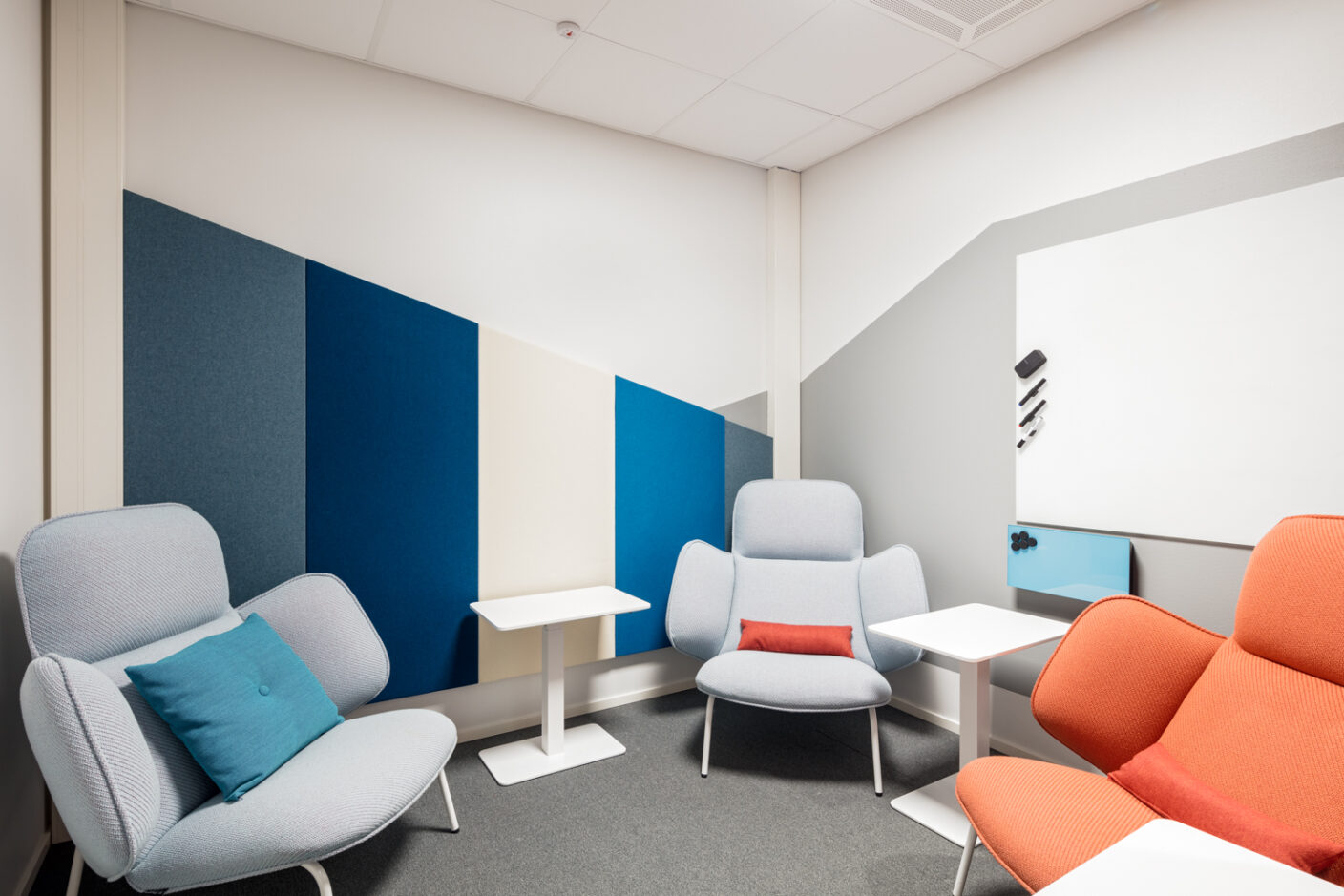Modern office space with three armchairs, two grey and one orange. The wall has a geometric design in shades of blue, white and grey. The space also has small white tables and decorative cushions.