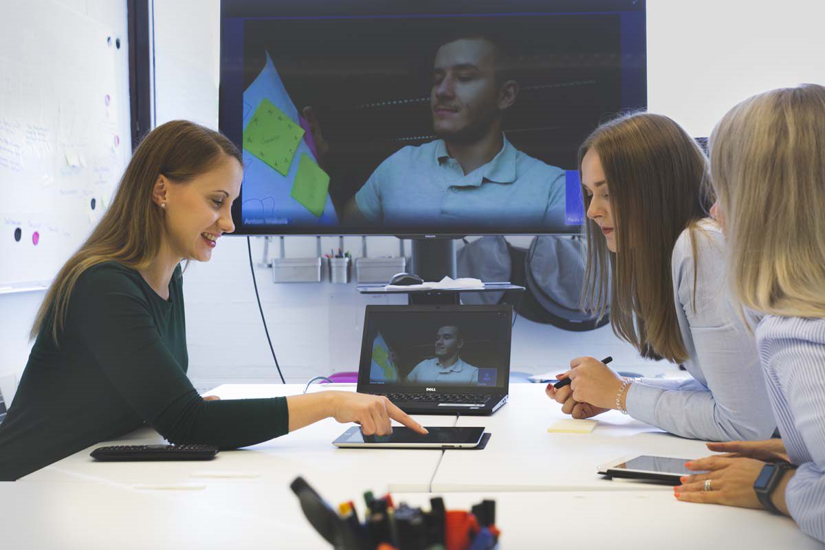 Kolme naista istuu neuvottelupöydän ympärillä ja katselee tablettilaitetta. Taustalla näkyy suuri näyttö ja kannettava tietokone, joissa molemmissa näkyy videopuhelun osallistuja.