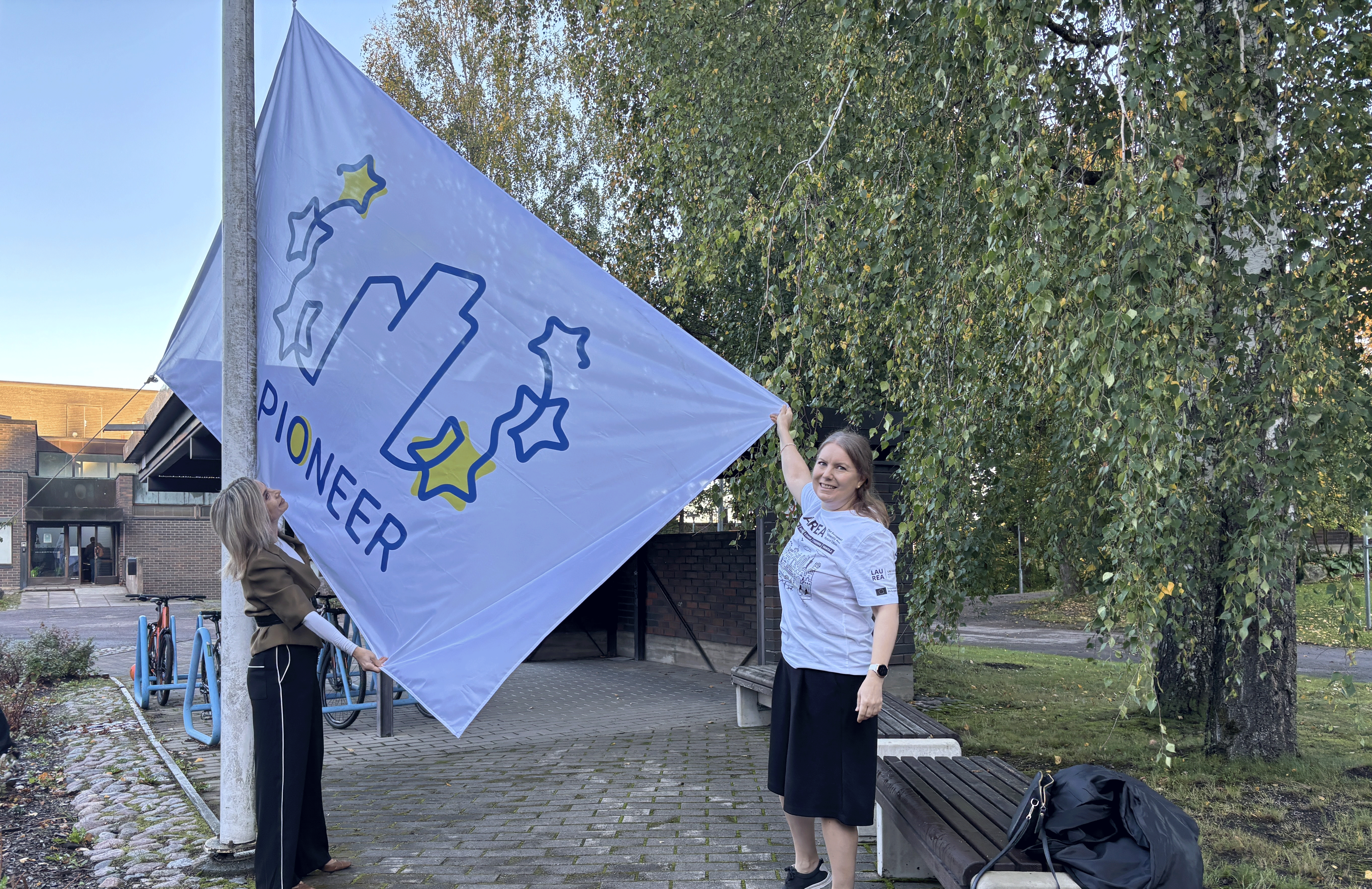 Two people raising a flag on a flagpole outside. The flag has the word 'PIONEER' and stars on it.