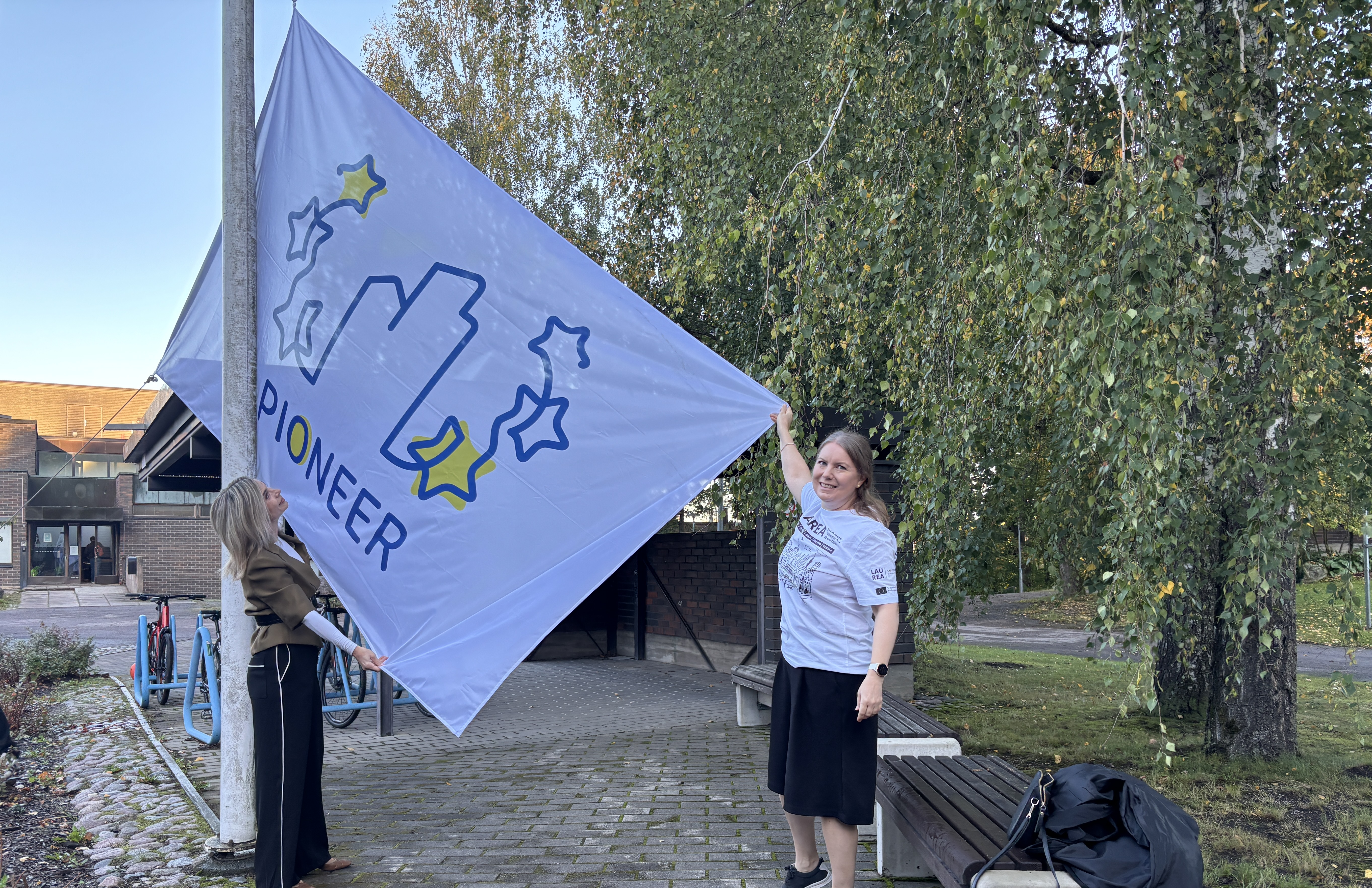 Kaksi henkilöä nostaa valkoisen PIONEER-lipun lipputankoon. Taustalla on puita ja rakennuksia.